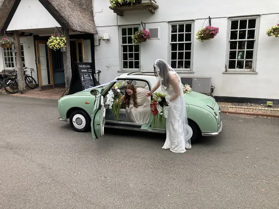 Bride at the door of a Nissan Figaro wedding car in Grantchester near Cambridge