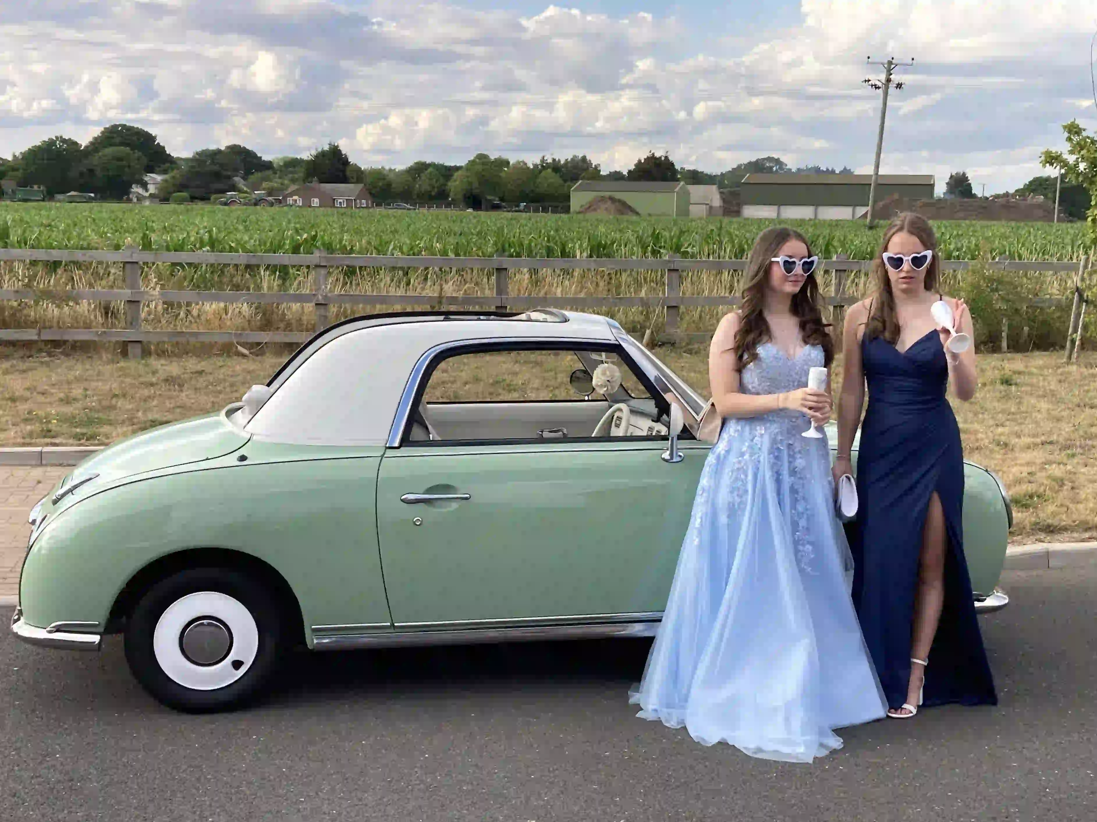 Two girls in prom dresses beside a Nissan Figaro prom car before a prom at Colchester's football stadium