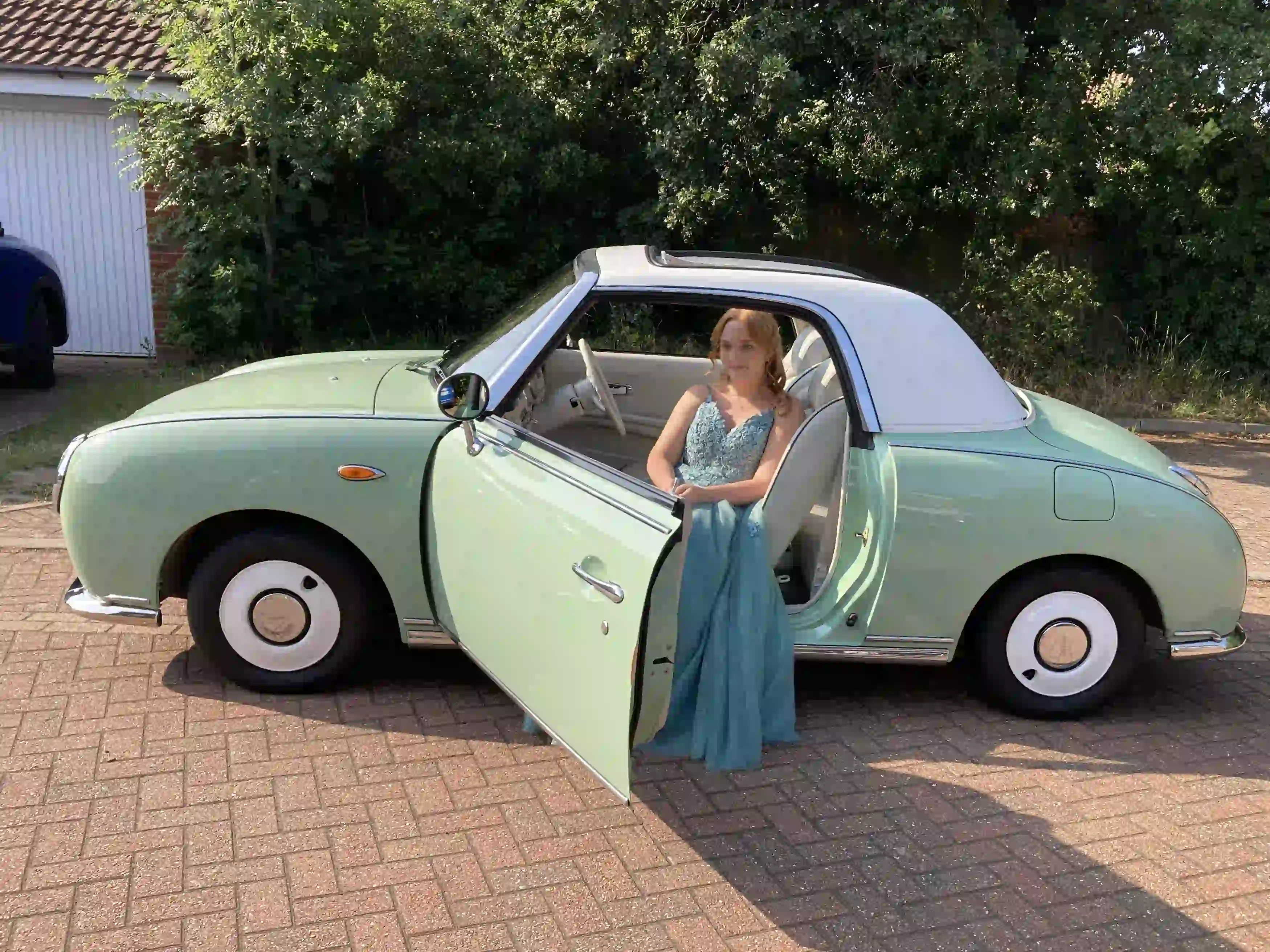 Hollie in her prom dress sitting in the passenger seat of a Nissan Figaro prom car with the door open, before her Norwich prom