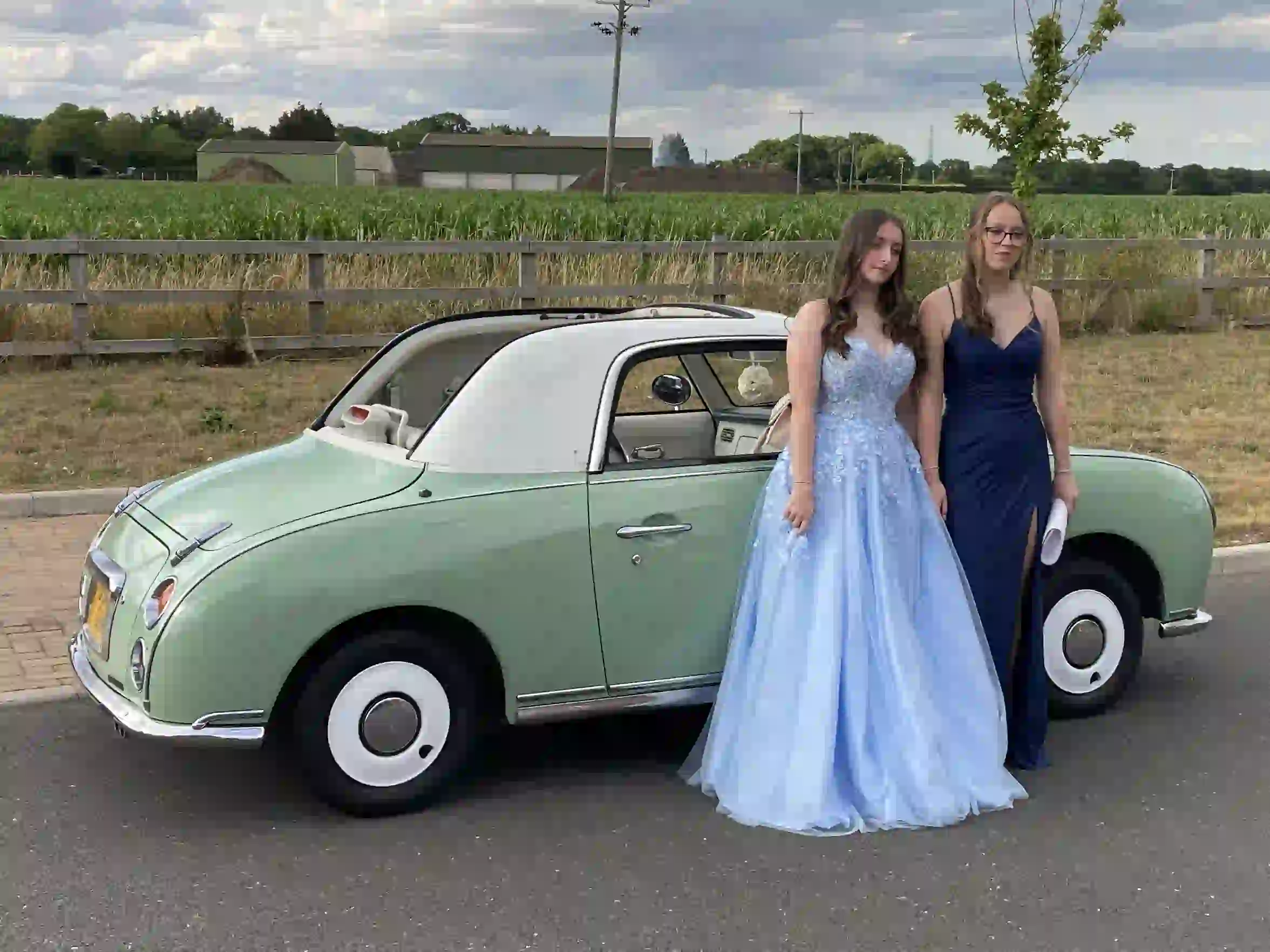 Two girls in prom dresses standing in front of a Nissan Figaro prom car on an Essex lane before their Colchester prom