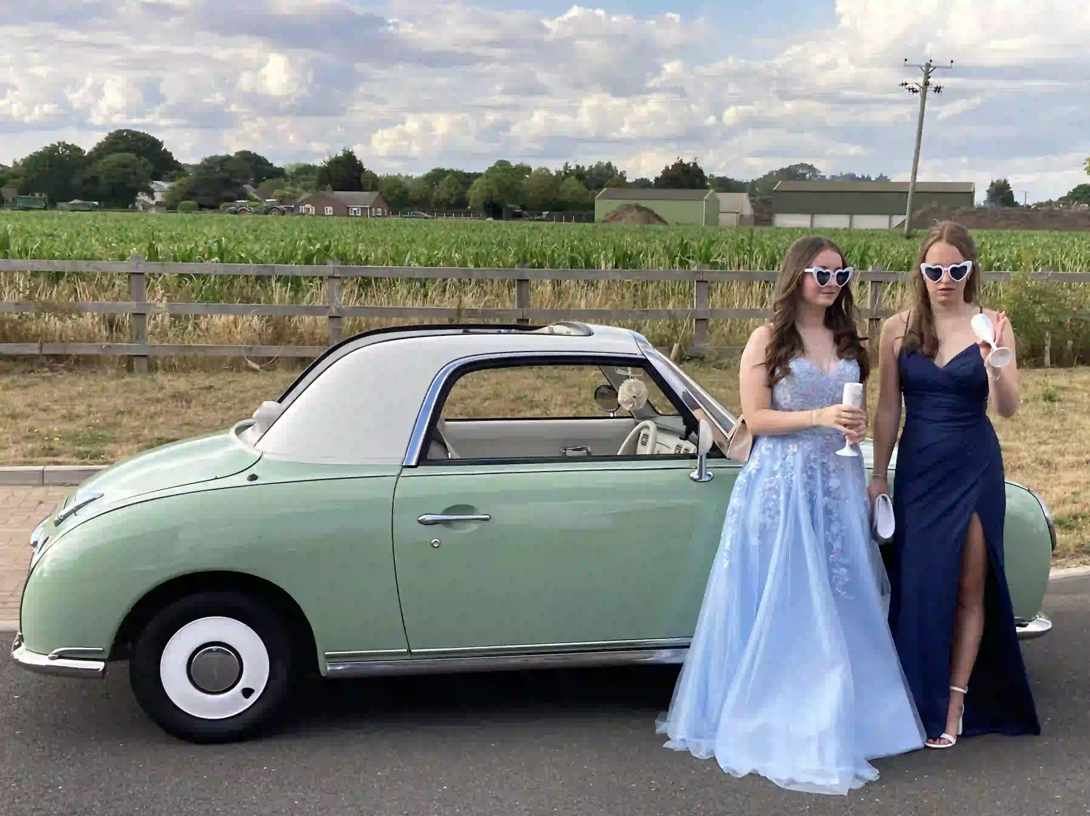 Two girls in prom dresses wearing novelty sunglasses and holding mock champagne glasses beside a Nissan Figaro prom car