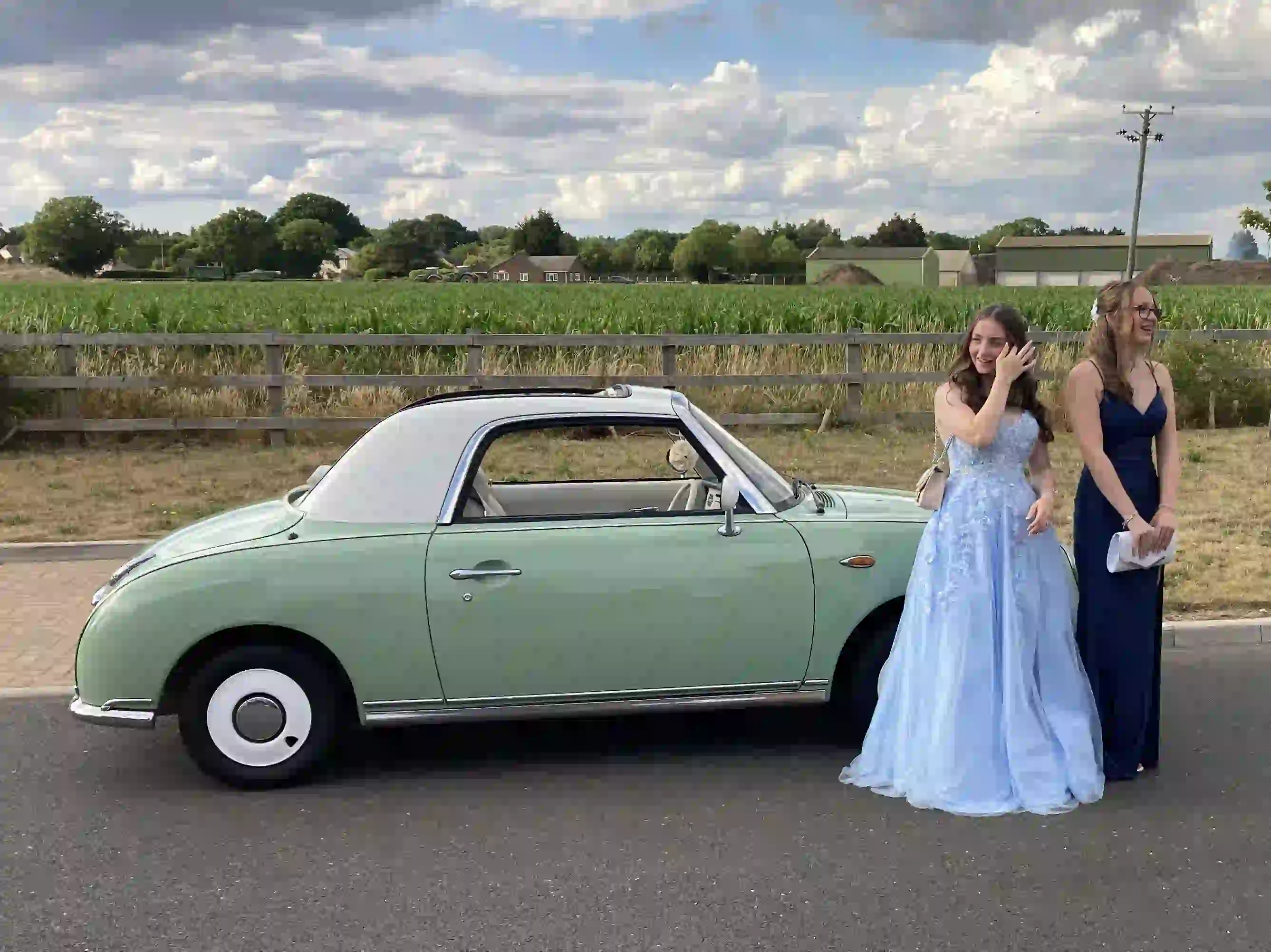 Two girls in prom dresses laughing in front of a Nissan Figaro prom car before their Colchester prom