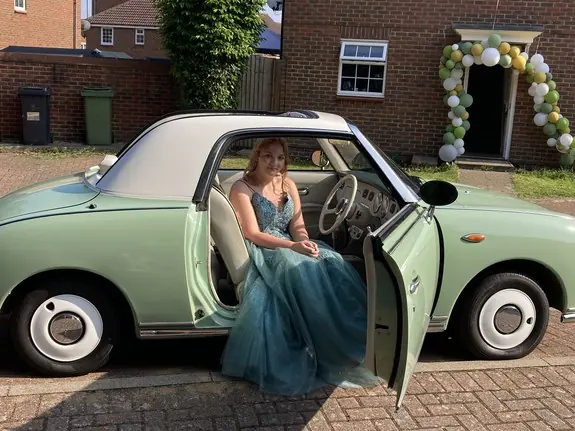 Girl in a prom dress stepping into a Nissan Figaro prom car outside a house