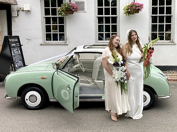 Two brides with their bouquets beside a Nissan Figaro wedding car in Cambridge