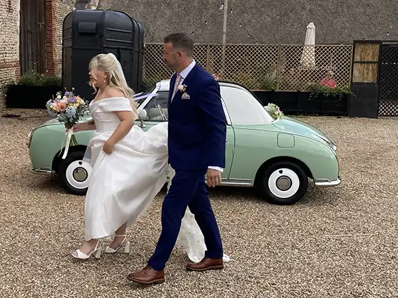 Bride and groom walking past a Nissan Figaro wedding car at Waxham Barn, Norfolk